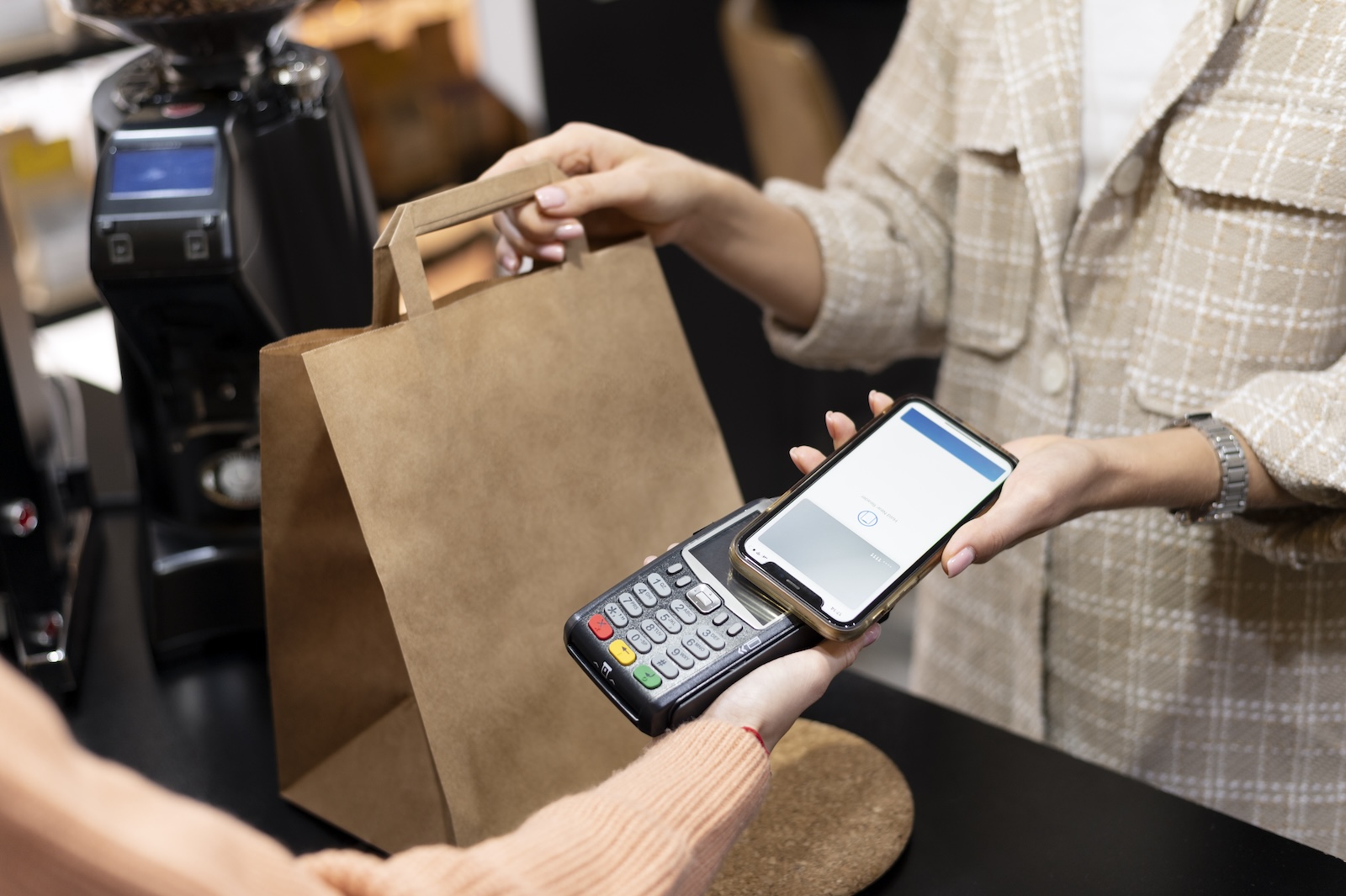 Imagem aproximada de uma pessoa realizando pagamento em uma maquininha conectada ao TEF com carteira digital no celular enquanto retira uma sacola.